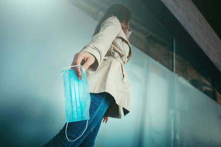 Cute girl is holding a medical mask in her hand and walking inside a modern building during pandemic of Coronavirus Covid-19の写真素材