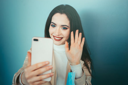 Cute girl is holding a medical mask in her hand and videocalling with smartphone during pandemic of Coronavirus Covid-19の写真素材