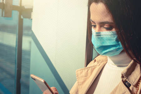 Girl with medical mask is using her smartphone in a modern building during pandemic of Coronavirus Covid-19の写真素材