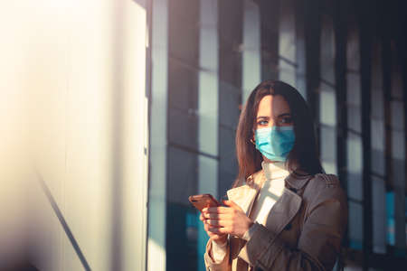Girl with medical mask is using her smartphone in a modern building during pandemic of Coronavirus Covid-19の写真素材
