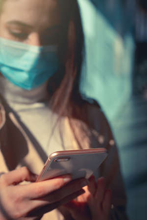 Girl with medical mask is using her smartphone in a modern building during pandemic of Coronavirus Covid-19の写真素材