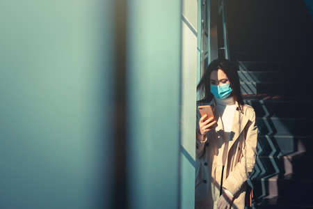 Girl with medical mask is using her smartphone in a modern building during pandemic of Coronavirus Covid-19の写真素材