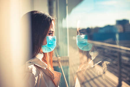 Girl with medical mask is looking out of the window during pandemic of Coronavirus Covid-19の写真素材