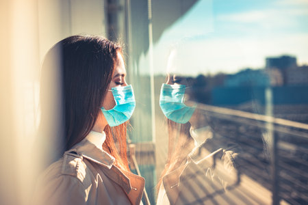 Girl with medical mask is looking out of the window during pandemic of Coronavirus Covid-19の写真素材