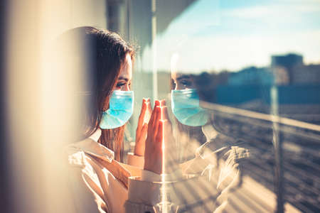 Girl with medical mask is looking out of the window during pandemic of Coronavirus Covid-19の写真素材