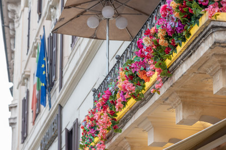 Ancient Italian Balcony Covered by Flowers and the European Flag Exposed on Blurred Backgroundの写真素材