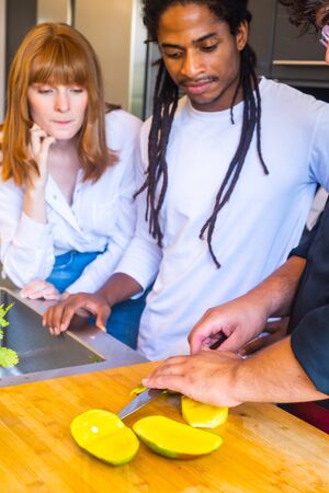 Stock vertical photo of a cook cutting a mango into pieces with a knife in front of a multicultural couple in a kitchenの写真素材