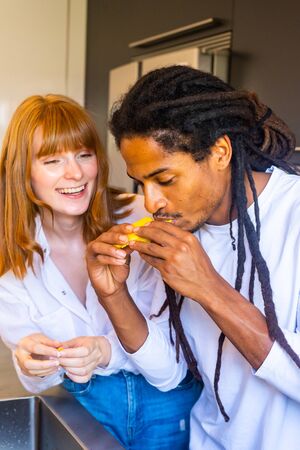 Stock vertical photo of a black guy with dreadlocks eating a mango in the kitchen with a young woman by his side laughing.の写真素材