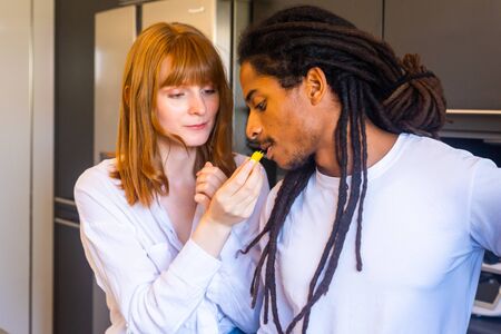 Stock photo of a young woman giving a piece of mango to a black boy with dreadlocks in the kitchen.の写真素材