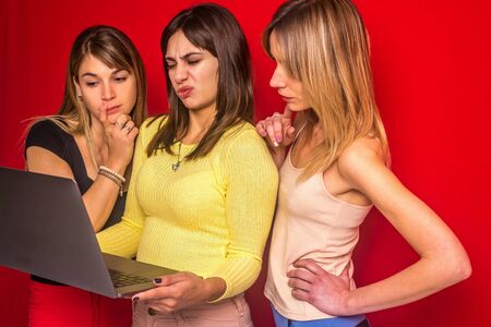 Stock photo of three girls standing with a laptop and a disgusting expression on a red backgroundの写真素材