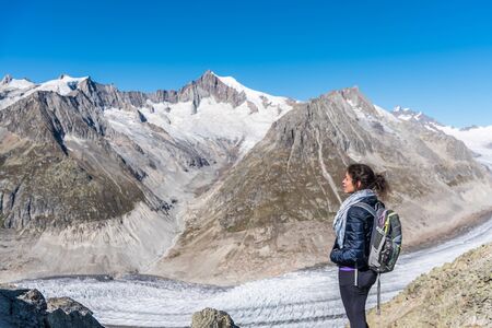 Young Caucasian woman with a backpack on her back looking at the monumental Aletsch Glacier.This...