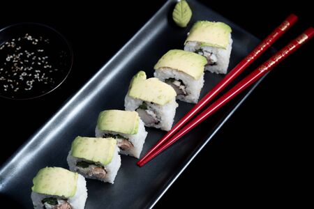 Sushi roll with avocado wrap, grilled salmon, Philadelphia cheese and green onions on black plate and red chopsticks. Small pot with soy sauce. Black background.の写真素材