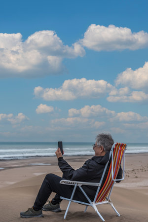 Mature adult male sitting on a beach chair on top of a dune facing the sea talking on video with...