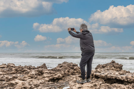 Mature adult man with grey hair and glasses taking a photo with his mobile standing on the rocks...