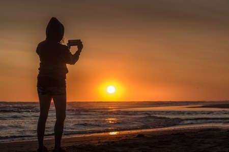 Silhouette of a hoodie woman on the beach taking a photo with her smartphone at sunset with the sun...