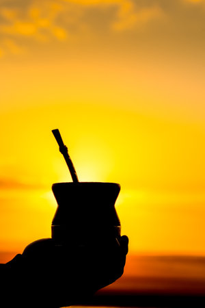 Vertical photograph of silhouette of a hand holding yerba mate drink at golden sunset. South...