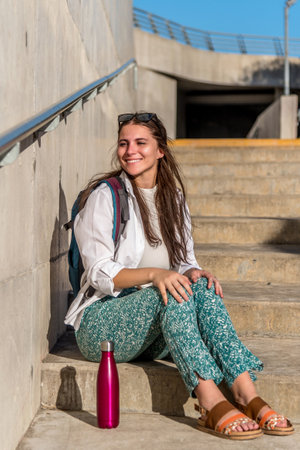 Cheerful tourist woman with backpack smiling while relaxing sitting outdoors on stairs.