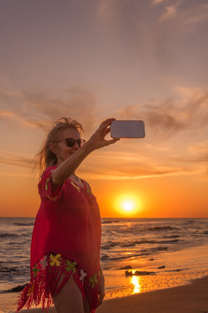 Senior woman with sunglasses taking a selfie with a mobile phone during sunset at the beach. Summer concept.の写真素材