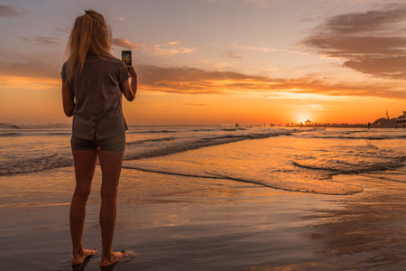 Woman using her mobile phone while taking photos of the sunset on the beach.の写真素材