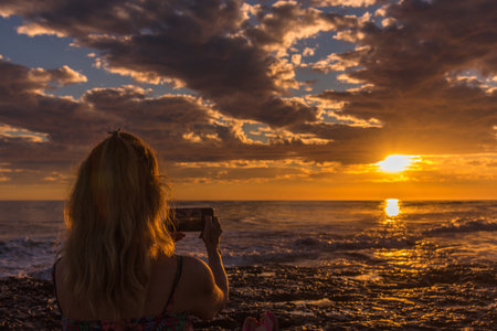 Woman taking photos of the sunset on the beach with mobile phone. Summer, travel and technology concept.の写真素材