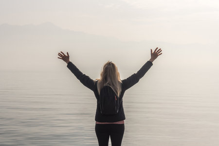 Traveler woman with arms raised standing in front of a lake in a misty day.