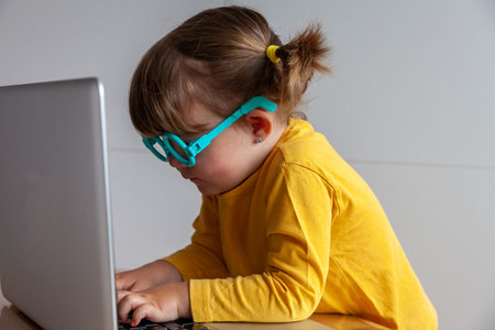Real Little girl in yellow t-shirt and blue glasses using laptop at homeの写真素材