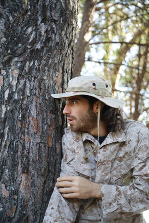Scared Soldier Hides Behind The Trunk Of A Tree In The Mountains From The Enemy