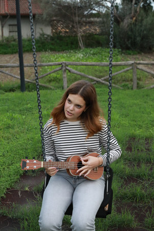 Young Woman Practices Playing The Ukulele Seated In A Swing