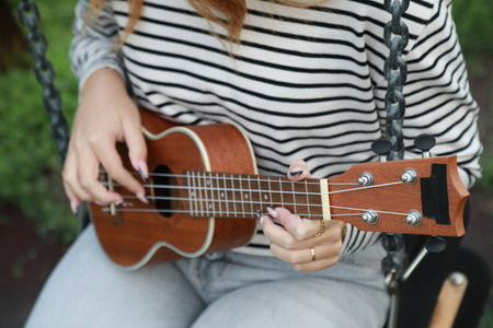 Young Woman Practices Playing The Ukulele Seated In A Swingの写真素材