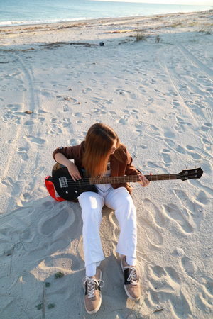 Girl Plays The Bass Instrument Sitting On The Beach At Sunset Near The Ocean