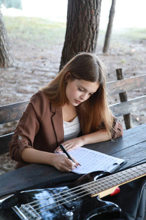 Girl Writes Music Notes On Sheet Music Sitting At The Park Table In The Natureの写真素材