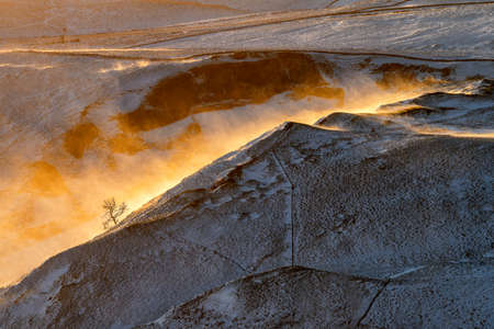 Strong wind blowing snow over ridge of a hill in the Peak District with golden morning light and...