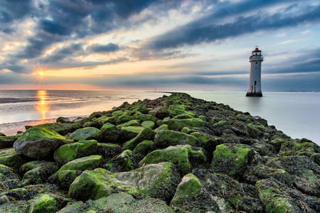 Lighthouse with moody dramatic clouds at sunset with algae rocks. New Brighton, Merseyside, UK.