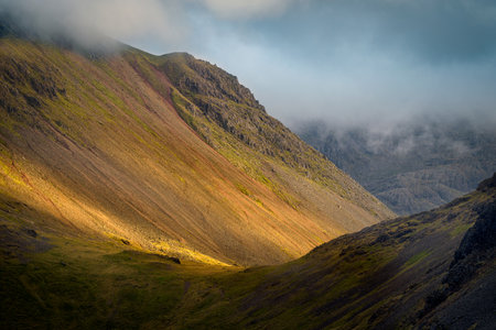 Broken Light Shining Onto Slopes Of Mountain Range With Dark And Dramatic Cloud. Lake District, UK.の写真素材