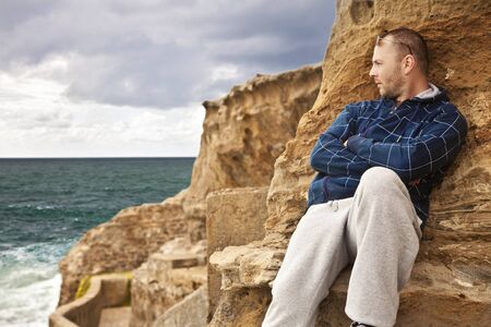 man laying on the rocks on the beach looking toward the sunlit water pensivelyの写真素材