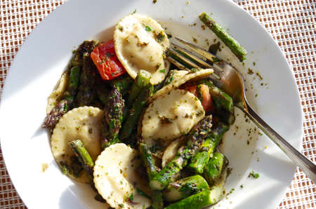 A white bowl of ravioli pasta with tomatoes, pesto and green asparagus and a fork on a white place mat.の写真素材