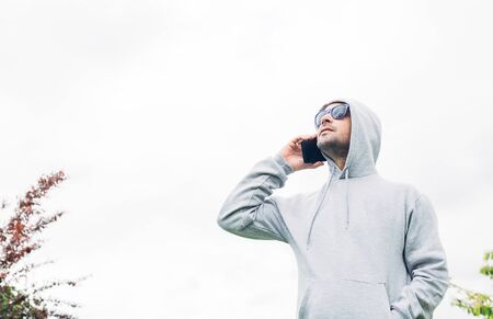 Young man in gray sweatshirt and blue sunglasses holding cell phone in his hands and smiling on a white backgroundの写真素材