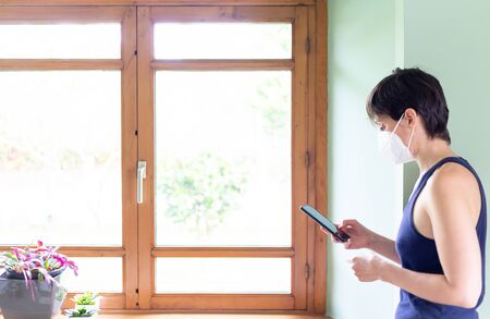 Woman stands by the window and wears a mask consulting the news about the Coronavirusの写真素材