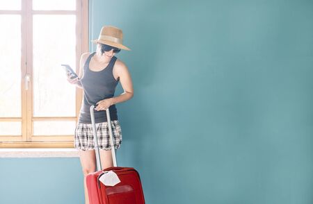 Woman in skirt, with straw hat and striped shirt using her mobile prepared with a red suitcase and her face mask for her vacation in Covids time.の写真素材
