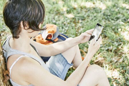 Girl sitting on the lawn checking her phone. Next to her is her backpack and her capの写真素材