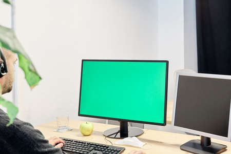 Man at his workplace with a computer and a green screen, mask, apple and glass of water on his desktopの写真素材