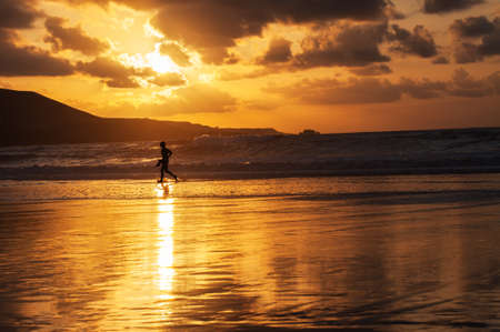 Silhouette of a surfer leaving the water with fins in hand and the sunset behind himの写真素材