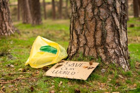 View of a cardboard banner on the ground with an environmental message next to a plastic bag at the foot of a tree. Concept of sustainability.の写真素材