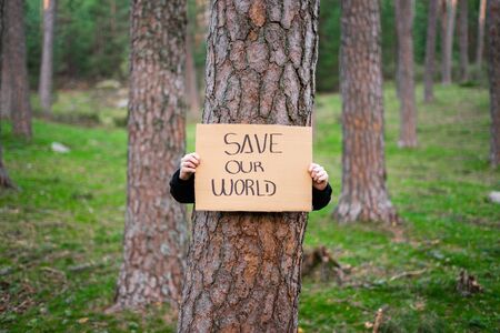 Close up view of teenage activist holding banner around a tree with environmental message. in the forest. Save our world. Concept of sustainability.の写真素材