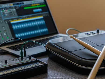 Close up of an electronic battery with drumsticks, an fm synthesizer and a laptop with a music...