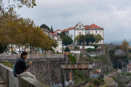 Two man somoking, sitting on the edge of a wall, while sightseeing. Buildings and trees on the background. Portoのeditorial素材