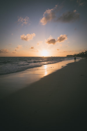 At dusk, a quiet beach scene unfolds with the sun setting over the ocean. Soft waves lap at the shore as silhouettes stroll along the water's edge, enjoying the tranquil beauty.の写真素材