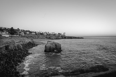 Sunset casts a calm glow over the ocean, with rocky cliffs and homes lining the coast. Seagulls fly...