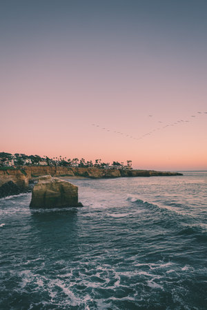 Waves gently lap against coastal rocks as a colorful sunset paints the sky. Birds fly in formation above the water, creating a serene atmosphere.の写真素材