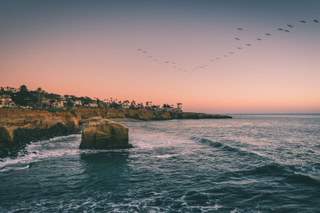 Waves gently crash against rocks as birds fly in a line across a colorful sunset sky near the coastline.の写真素材